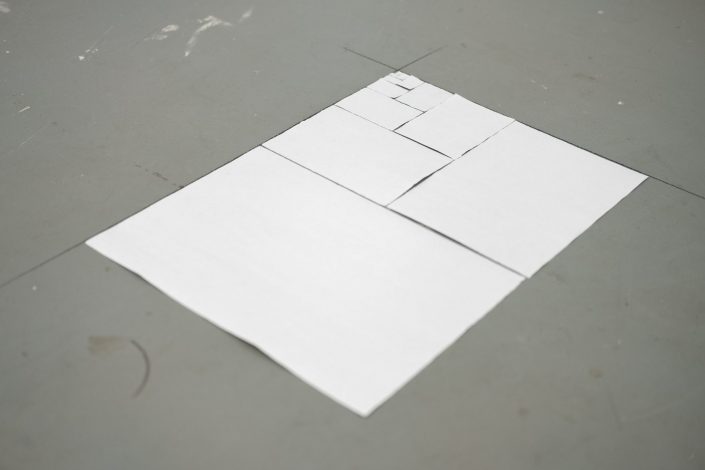 Installation of standard sized paper folded in half, torn and placed on a marked floor to create the interlocking pattern of the ISO 216 paper series as it diminishes in size.