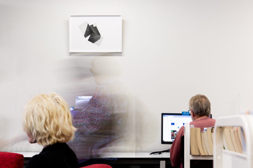 People seated in a library working on computers with the blurred image of one person walking past two black and white photo collages made from images of an empty room